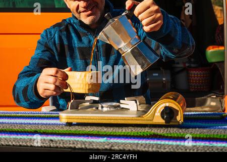 Ritaglia un anonimo maschio sorridente con abiti informali versando il caffè in una tazza mentre sei seduto al tavolo con una stufa a gas portatile in camper nel canyon Ro Foto Stock