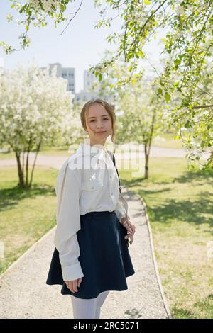 Una giovane adolescente studentessa sorridente con la borsa della scuola a spalla che guarda la fotocamera mentre si trova sul sentiero nel parco verde del campus con edifici Foto Stock