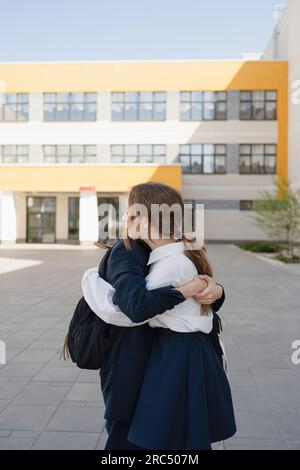 Giovane studentessa adolescente che abbraccia un compagno di classe anonimo in uniforme con lo zaino e guarda lontano mentre si trova nel campus scolastico alla luce del giorno Foto Stock