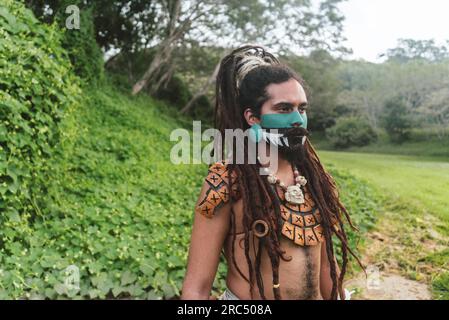 Ritratto di un guerriero maschio barbuto con viso dipinto e capelli intrecciati indossando costumi da collo mentre si è in piedi nel verde parco con alberi e guardando lontano Foto Stock