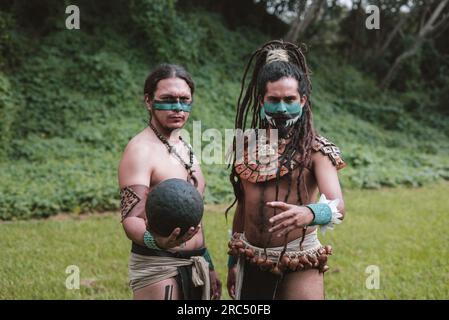 Gruppo di guerrieri maschi etnici maya con accessori tradizionali e pittura del viso che guardano la fotocamera mentre si trovano in piedi con la palla nera stampata in palla erbosa Foto Stock