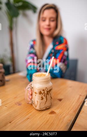 Attenzione selettiva alla bottiglia di delizioso caffè cremoso con sfondo sfocato di giovani donne in abiti informali seduti a un tavolo di legno nel ristorante Foto Stock