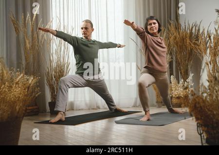 Corpo completo di marito e moglie in abbigliamento sportivo che si allunga in Virabhadrasana yoga posa mentre si pratica yoga insieme sul tappetino a casa Foto Stock