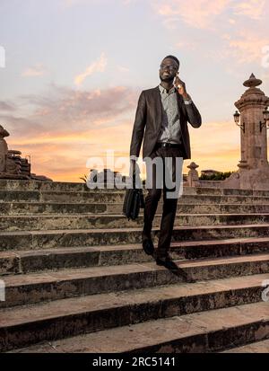 Corpo pieno di uomini afro-americani sorridenti in abito formale alla moda con valigetta e occhiali al telefono mentre cammini sulle scale nella città vecchia di ag Foto Stock