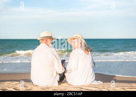 Vista posteriore di un uomo e una donna anziani amorevoli con cappelli di paglia seduti insieme sulla spiaggia e guardarsi l'un l'altro Foto Stock