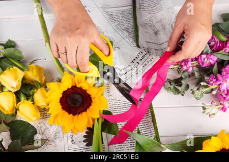 Fiorista anonimo con forbici che taglia l'arco rosa e organizza il bouquet di girasole, rose gialle e fiori viola al tavolo wi Foto Stock