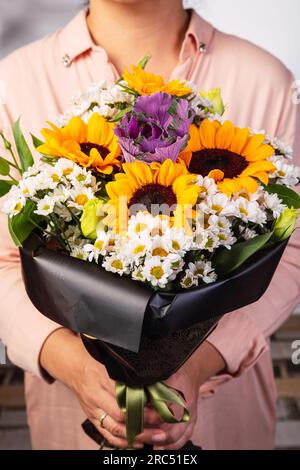Coltiva una fiorista femminile irriconoscibile in abiti eleganti e casual, con un mazzo di fiori freschi e aromatici in una confezione regalo avvolta mentre sei in piedi alla luce del giorno Foto Stock
