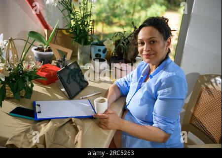 Designer floreale donna incinta che sorseggia un caffè, sorride guardando la macchina fotografica mentre fai una pausa caffè in uno studio floreale Foto Stock