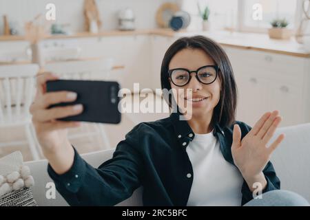 Una donna sorridente risponde alle videochiamate, salutando, seduto sul divano. Giovani blogger chatta online, accoglie i follower. Foto Stock