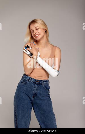 Studio ritratto a mezza lunghezza di una bella ragazza bionda con disabilità che indossa un braccio prostetico bionico sensoriale. Donna vestita con jeans blu e beig Foto Stock