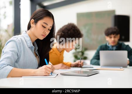 Signora asiatica seduta alla scrivania e prendendo appunti, scrivendo saggi, studiando per l'assegnazione della classe con compagni di classe sullo sfondo Foto Stock