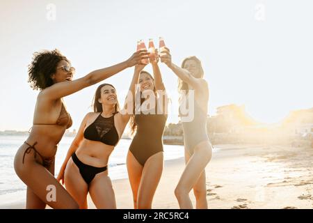 Brindisi all'amicizia. Allegri e diversificate giovani donne in costume da bagno che alzano le bottiglie di birra mentre si trovano in spiaggia Foto Stock