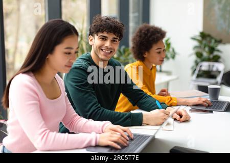 Gruppo di diversi studenti internazionali seduti alla scrivania in uno spazio di coworking, concentrati sul sorriso che scrive in un copybook Foto Stock