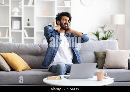 Maschio indiano stanco che massaggia il collo infiammato dopo aver lavorato sul laptop a casa Foto Stock