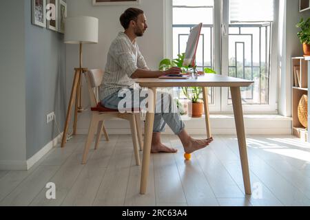 Uomo freelance che lavora al computer a casa facendo esercizio di recupero del piede usando la palla di silicone massaggiante. Foto Stock