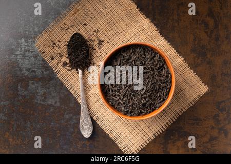 Asciuga le foglie di tè nero in un recipiente di argilla e un cucchiaio d'annata su uno sfondo scuro vista dall'alto. Tè nero a foglie grandi e a foglie piccole. Foto Stock