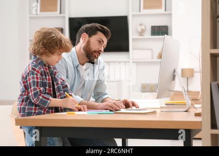 Uomo che lavora a distanza a casa. Un padre impegnato a parlare con lo smartphone mentre suo figlio sta disegnando alla scrivania Foto Stock