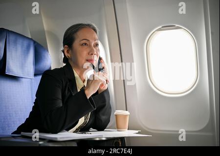 Una donna d'affari asiatica premurosa e seria o un CEO donna in una tuta formale è al telefono con il suo cliente d'affari durante il volo Foto Stock