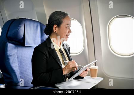 Una donna d'affari asiatica anziana felice e sorridente che utilizza il suo tablet digitale durante il volo. Business people e concetto di trasporto Foto Stock
