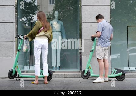 Scooter elettrici condivisi a noleggio di fronte alla vetrina di un negozio di moda a Vilnius. Una coppia decide di affittarli. Mobilità elettronica Foto Stock