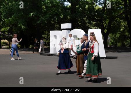 I nazionalisti lettoni in costumi nazionali sventolano bandiere nel centro di riga, in Lettonia. Nome della città RIGA in grandi maiuscole bianche Foto Stock