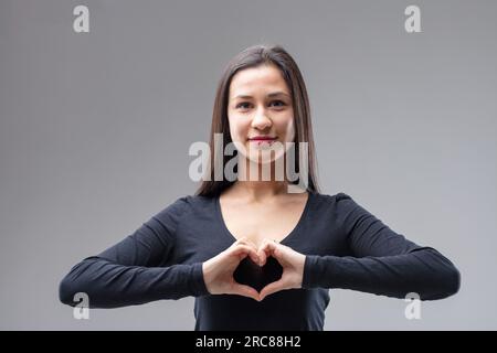 Ritratto frontale a metà busto di una giovane donna che forma un cuore con le dita. Ti amo! Mi piace tutto questo! Adoro questa situazione o cose del genere." Si mette Foto Stock