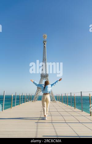 Grande modello della Torre Eiffel sulla spiaggia. Una donna cammina lungo il molo verso la torre, indossando una giacca blu e jeans bianchi. Foto Stock