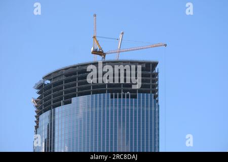 La gru a torre gialla sulla sezione superiore è la costruzione di un moderno e alto grattacielo contro il cielo blu senza nuvole Foto Stock