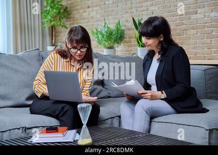 Due donne mature d'affari sedute sul divano in ufficio, a parlare, a parlare di lavoro Foto Stock