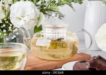 Tè estetico, teiera, tè verde e cioccolato in interni biofilici tra fiori bianchi. Stile di vita femminile e tranquillo, accogliente concetto di casa. Foto Stock