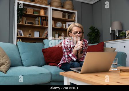 Stanco stressato turbato donna matura di mezza età che sente mal di testa dopo il lavoro al computer seduto a casa sul divano, triste stanco vecchia nonna anziana signora Foto Stock
