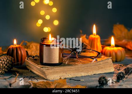 Candela Halloween STILL Life con cera di soia nel vaso di vetro ambrato scuro con etichetta vuota per il trucco. Candele a forma di zucca con coni, foglie, Old Book Foto Stock