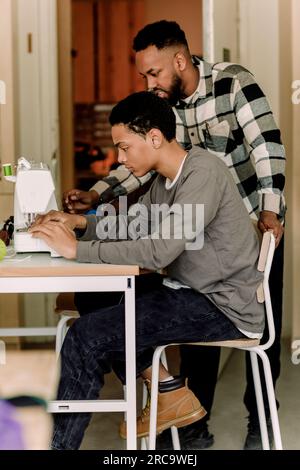 Ragazzo adolescente che impara a cucire usando la macchina da cucire da un insegnante in classe d'arte alle superiori Foto Stock