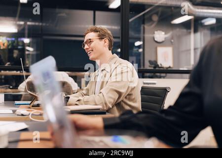 Un uomo d'affari felice che indossa occhiali da vista seduto alla scrivania che lavora in ufficio Foto Stock