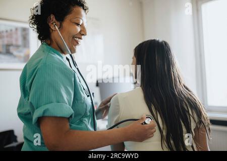 Vista laterale di una donna sorridente che esamina una giovane donna con stetoscopio in clinica Foto Stock