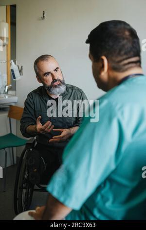 Paziente maturo in sedia a rotelle che spiega il medico in clinica Foto Stock