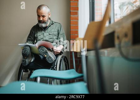 Uomo maturo con disabilità che legge riviste mentre è seduto in sedia a rotelle in sala d'attesa Foto Stock