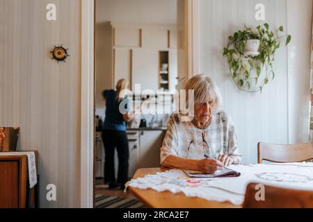 Donna anziana che fa rompicapo nel libro mentre si siede al tavolo da pranzo a casa Foto Stock