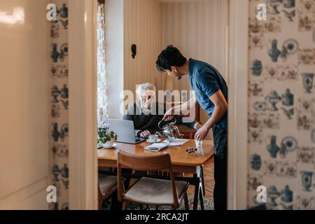 Vista laterale del lavoratore sanitario maschile che serve il tè all'uomo anziano a casa Foto Stock