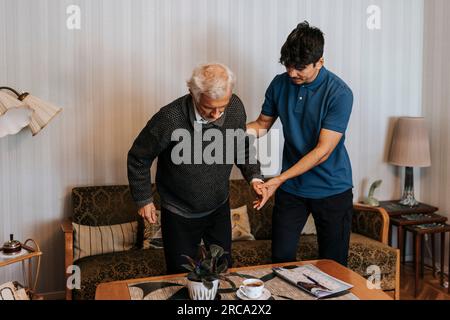 Assistente di cura maschile che sostiene l'anziano mentre si alza dal divano a casa Foto Stock