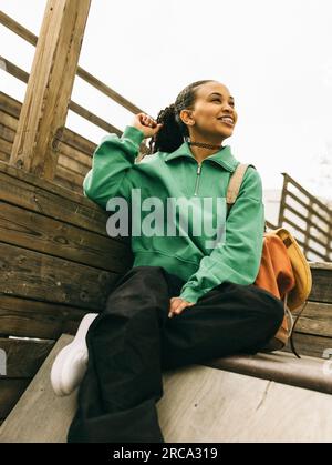 Giovane donna sorridente che guarda lontano mentre si siede sui gradini Foto Stock