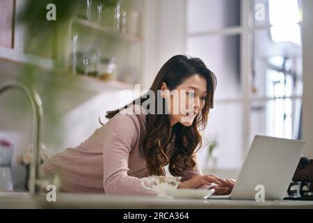 giovane donna d'affari asiatica che lavora in cucina a casa utilizzando un computer portatile Foto Stock