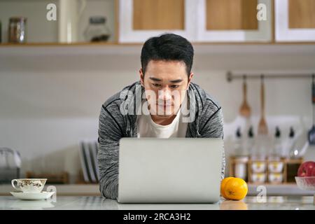 giovane uomo d'affari asiatico che lavora in cucina a casa usando un computer portatile Foto Stock