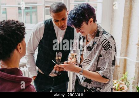 Persona non binaria che condivide lo smartphone con un uomo d'affari che tiene un bicchiere durante l'evento al centro convegni Foto Stock