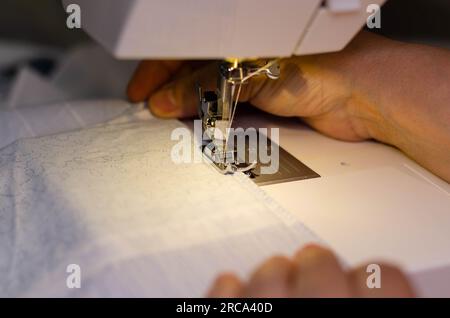 Primo piano delle mani di una sarta che cuce tessuto bianco su una macchina da cucire Foto Stock