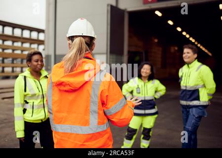 Vista posteriore di una donna ingegnere in abbigliamento da lavoro protettivo che discute con i lavoratori in fabbrica Foto Stock