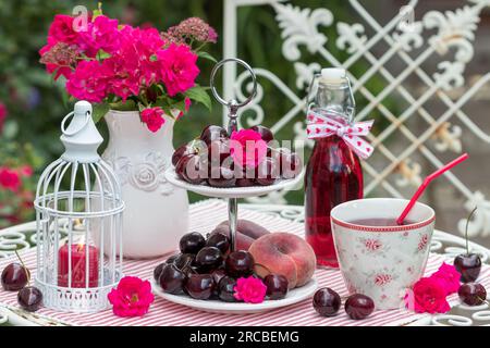 disposizione del tavolo con ciliegie dolci fresche e pesche su un supporto per torta a più livelli e succo d'uva in una tazza e una bottiglia di vetro Foto Stock