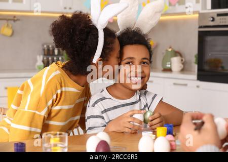 Felice madre afro-americana che bacia suo figlio carino mentre dipinge le uova di Pasqua al tavolo in cucina Foto Stock