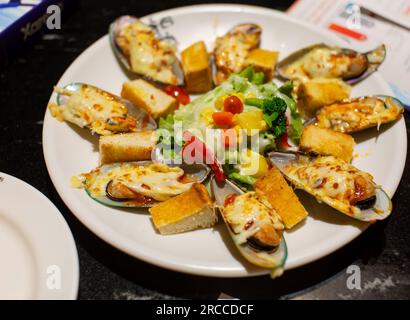 Otto fette di cozze neozelandesi cotte con formaggio su un piatto rotondo bianco e insalata verde vista ravvicinata dal lato basso concentrata sul cibo Foto Stock