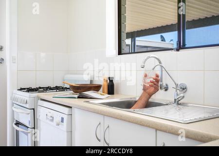 Mano che esce dal lavello riempiendo il vetro con acqua Foto Stock